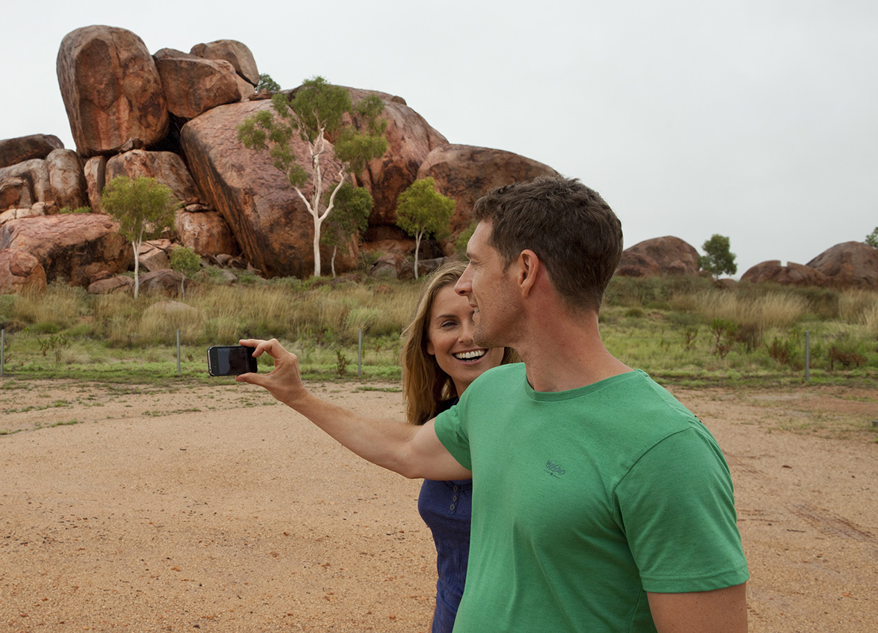 Lady and man standing infront of the Devils Marbles using a mobile phone.