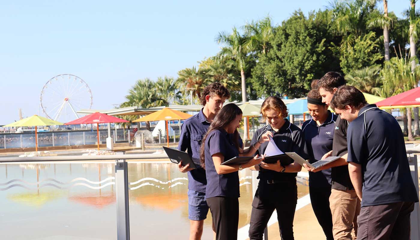 Students looking over some paperwork and laptops in an outdoor water park in the background